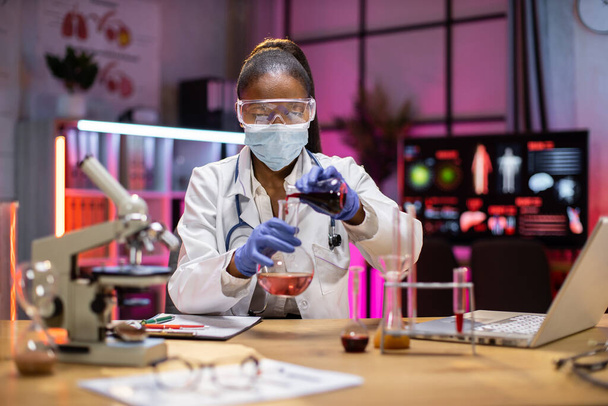 Medical research laboratory: portrait of a attractive female scientist using digital laptop computer, analysing liquid biochemicals in a laboratory flask. Advanced scientific biotechnology laboratory. - 写真・画像