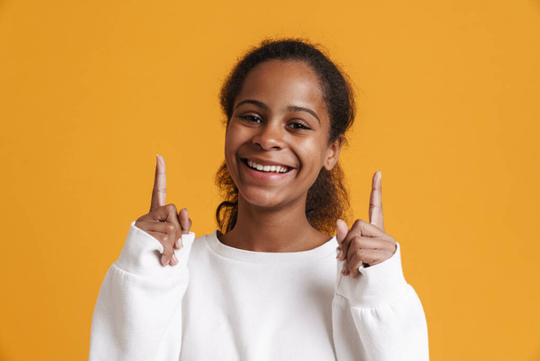 Brunette black girl smiling while pointing fingers upward isolated over yellow background - Foto, Imagem