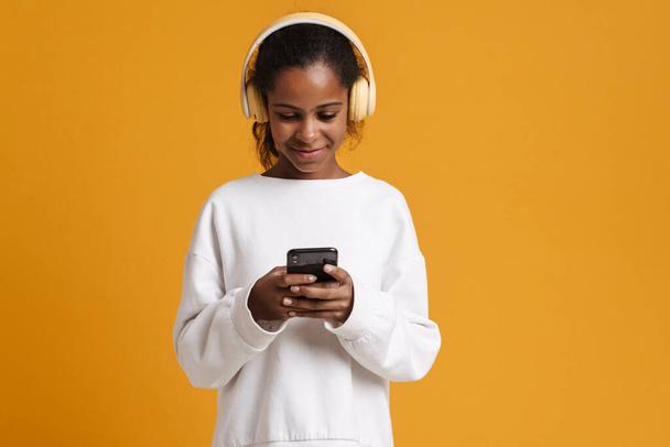 Brunette black girl in headphones smiling while using cellphone isolated over yellow background - Foto, immagini