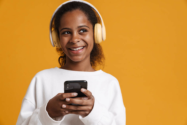 Brunette black girl in headphones smiling while using cellphone isolated over yellow background - Foto, afbeelding
