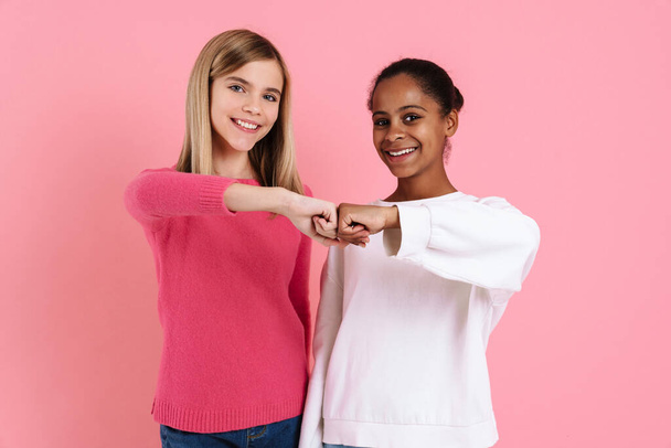 Multiracial two girls wearing sweaters smiling and fists bumping isolated over pink wall - Фото, зображення