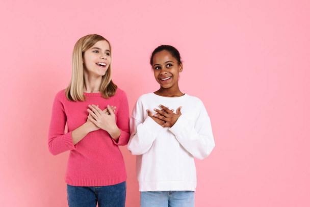 Multiracial two girls smiling while holding hands on their chests isolated over pink wall - Fotó, kép