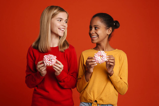 Multiracial two girls smiling while posing with donuts isolated over red wall - Foto, afbeelding