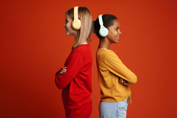 Multiracial two girls smiling while listening music with headphones isolated over red wall - Photo, Image
