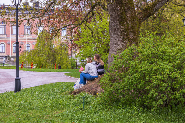 Beautiful view of two young woman sitting on roots of big old tree in park. Sweden. Uppsala. 05.14.2022. - Foto, Imagem