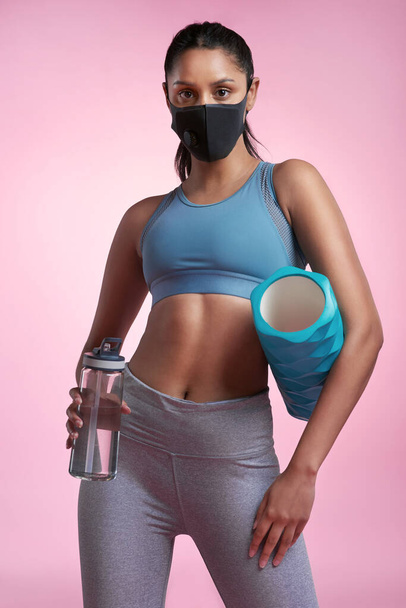 Quitting is never an option. Cropped portrait of an attractive and sporty young woman wearing a mask while posing with a foam roller and water bottle in studio against a pink background - Foto, imagen