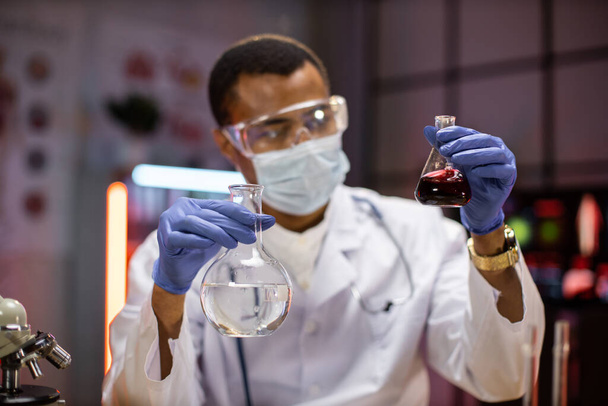 Scientific innovations. Positive smart enthusiastic african american man in protective mask and goggles scientist looking at the test tubes and smiling while developing a vaccine. - Foto, Bild