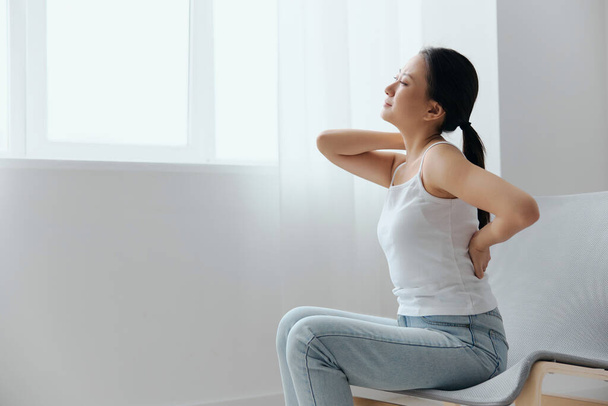 Back pain. Suffering from osteochondrosis after long study work pretty young Asian woman touching painful lower back at home interior living room. Injuries Poor health Illness concept. Cool offer - Photo, Image
