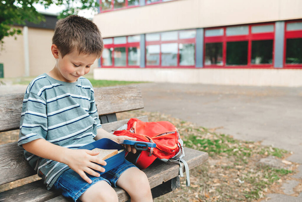 The boy sits on a bench in the school yard and takes out a sandwich from the lunch box for a snack. Nutrition for children while studying. - Foto, immagini