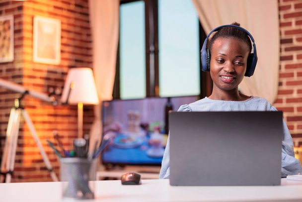 Young african american woman in headphones watching videos on laptop at home. Smiling female student attending remote university lesson, studyin with computer in modern room - 写真・画像