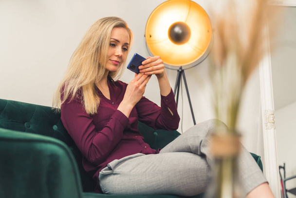 Pretty slim blondie camfortably sitting and intentively watching a video on her phone - studio room background. High quality photo - Foto, afbeelding