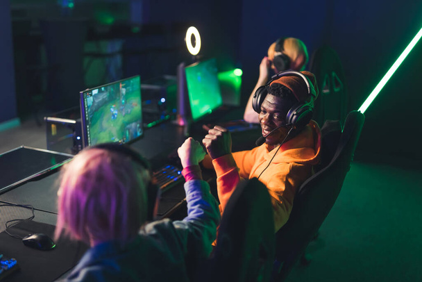 Teenage gamers wearing headsets fistbumping competing in an online gaming tournament. Professional gaming setup. Colored lights. High quality photo - Photo, Image