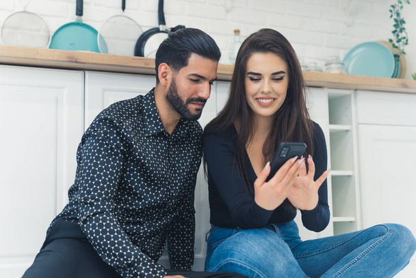 Man with dark hair and beard wearing patterned shirt looking at his girlfriend using smartphone and smiling sitting on the floor together. Young couple. Indoor shot. High quality photo - Foto, Imagem