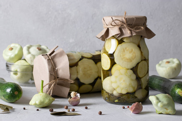 Pickled patissons and zucchini with dill, garlic and pepper in glass jars on light gray background - Photo, image