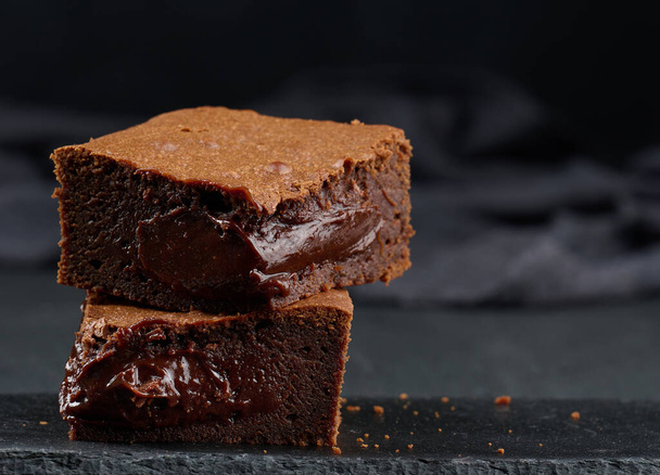 Baked pieces of chocolate brownie pie on black table, delicious dessert - Zdjęcie, obraz