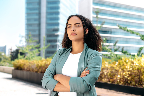 Portrait of a serious elegant confident young mixed race woman with curly hair, formally dressed, stands outdoors against the background of the business center, with arms crossed, looks at camera - Photo, image