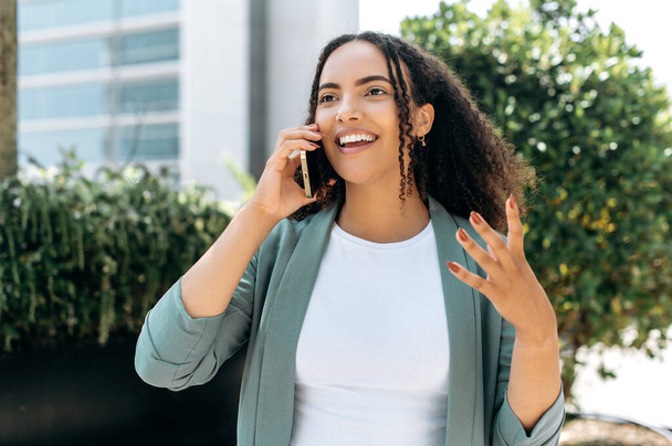 Beautiful positive confident mixed race young woman with curly hair, wearing formal clothes, emotionally talking on a smartphone, standing outdoors, gesturing with her hand, smiling friendly - Foto, imagen