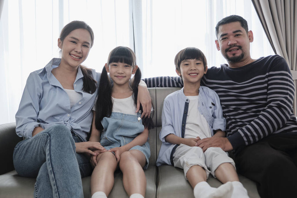 Portrait of Asian Thai family, adult dad, mum with little children happiness home living together, looking at camera, leisure on sofa in white room, lovely weekend and wellbeing domestic lifestyle. - Foto, Bild