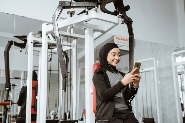 beautiful muslim sporty woman taking a break after exercising at the gym sitting on the floor and using phone - Foto, immagini