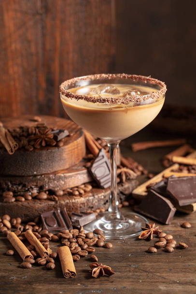 Irish cream and coffee cocktail in a glass with ice on an old wooden background. Coffee beans, cinnamon, anise, and pieces of chocolate are scattered on the table.  - Photo, Image