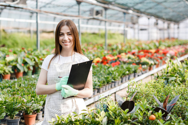 Retrato de mujer jardinero en el trabajo en invernadero con cuaderno examina las flores en crecimiento en invernadero. - Foto, imagen