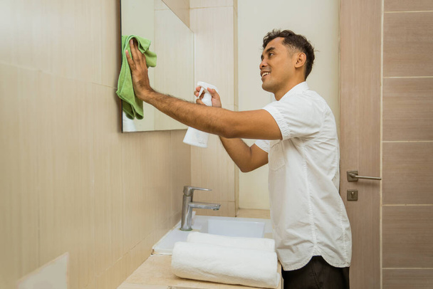asian male janitor spraying and wiping the mirror with rag in the bathroom - Photo, Image