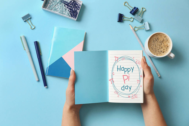 Female hands holding notebook with text HAPPY PI DAY, cup of coffee and stationery on light blue background - Foto, immagini