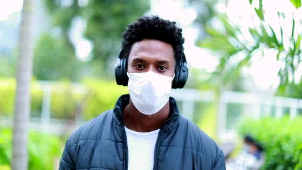 Young black man wearing surgical mask and headphones walking towards camera - Materiaali, video