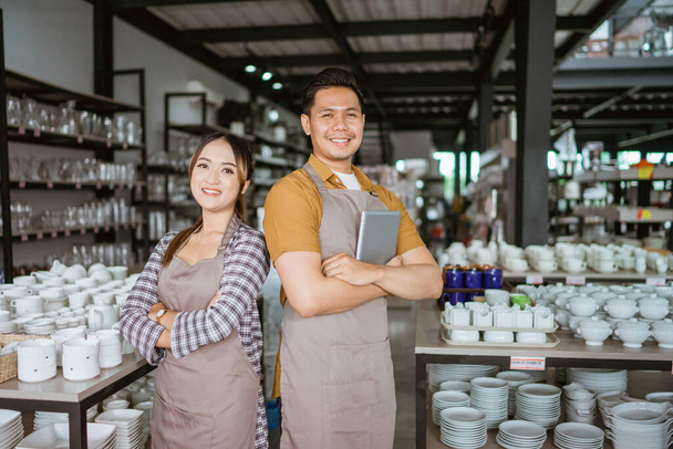 Two shop owners in apron holding digital tablet standing back to back in houseware store - Foto, Imagen