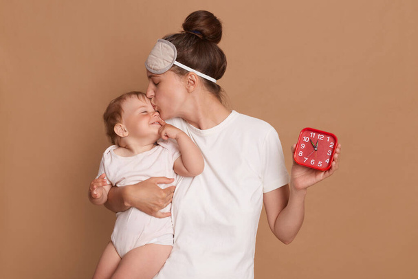 Horizontal shot of attractive woman wearing white t shirt and blindfold holding baby daughter and red alarm clock, mother kissing her cute child isolated over brown background. - Foto, afbeelding