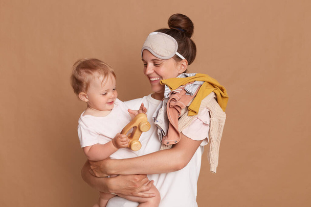 Portrait of beautiful young adult mother with a baby in her arms, woman wearing wearing white t shirt and white mask looking at her baby with smile, expressing happiness. - Photo, image