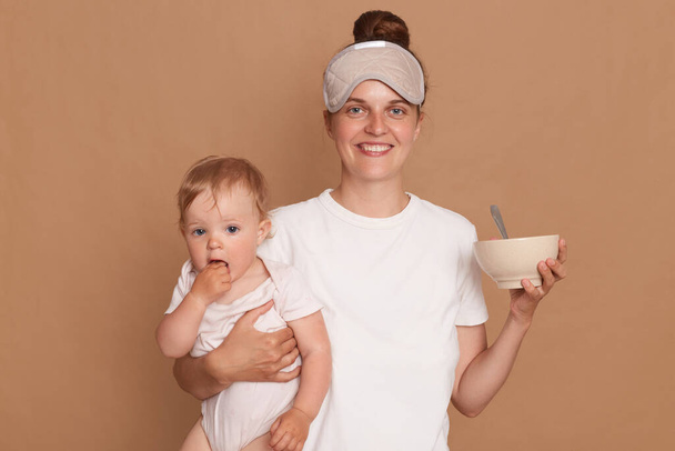 Horizontal shot of smiling happy positive mother with cute daughter, holding baby daughter in hands and plate with breakfast, looking at camera with toothy smile. - Photo, image