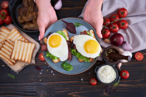 Fried eggs, bacon and cream cheese sandwiches on a plate on wooden table at domestic kitchen. - Photo, image