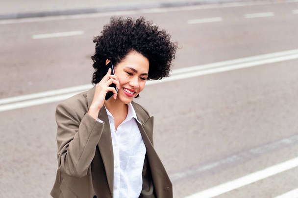 portrait of a smiling latin business woman walking by the street talking by phone, concept of urban lifestyle and success, copy space for text - Photo, image