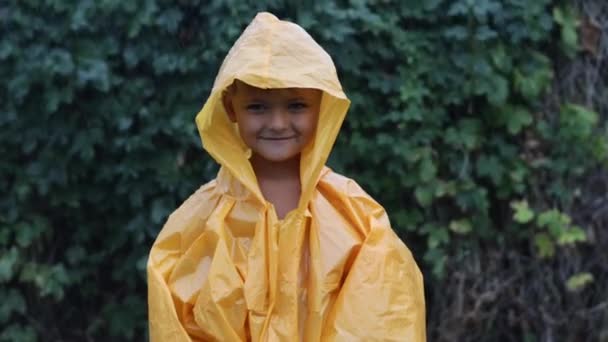 Een jongetje staat in de regen in een regenjas. Zomer regen druipt op de man in de regenjas. Kindervreugde over klimaatverandering.  - Video