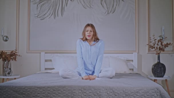 cheerful caucasian woman with freckles on young face yawning and stretching posing with crossed legs sitting on bed happy morning at home or hotel - Filmati, video