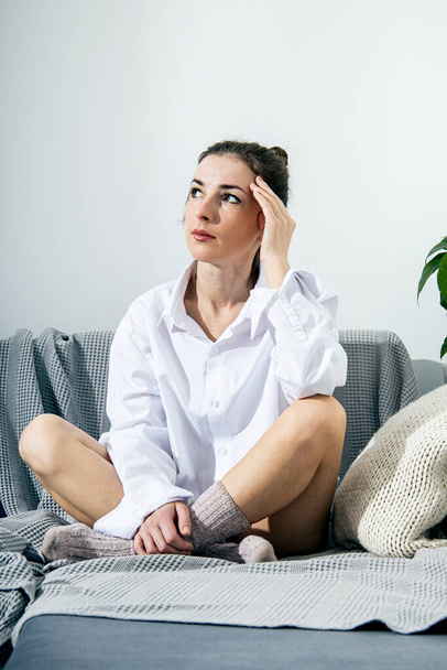 Thoughtful young woman in white shirt and socks sitting on sofa  - Photo, image