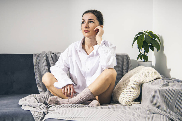 Thoughtful young woman in white shirt and socks sitting on sofa  - Фото, изображение