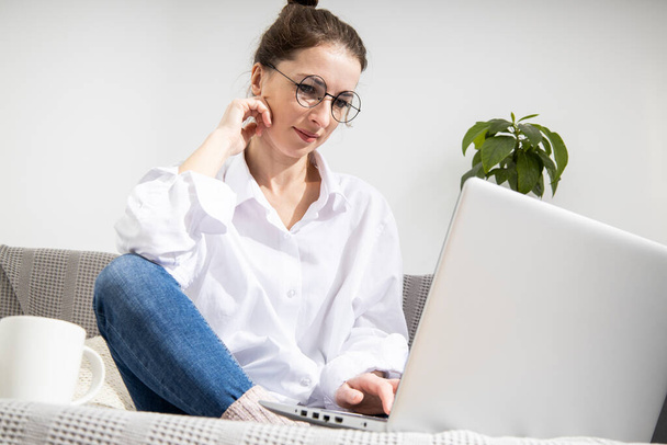 Jovem mulher em uma camisa branca com um laptop trabalhando enquanto se senta no sofá - Foto, Imagem