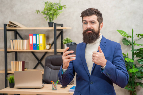 confused bearded businessman drinking takeaway coffee in the office show thumb up, coffee break. - Foto, afbeelding