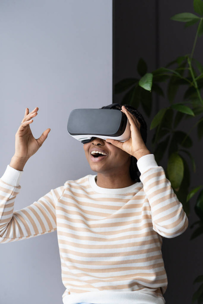 African american woman playing on vr glasses at home. Virtual reality and wearable tech concept with young female has fun wearing headset goggles. Digital technology for entertainment and business - Photo, Image