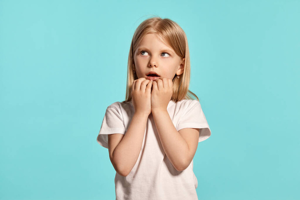 Close-up studio shot of a cute blonde kid with a long hair, in a white t-shirt, looking scared while posing over a blue background with copy space. Concept of a happy childhood. - Foto, imagen
