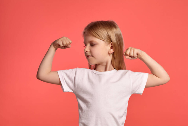 Close-up studio shot of a charming blonde little girl with a long hair, in a white t-shirt, showing her muscles while posing over a pink background with copy space. Concept of a joyful childhood. - Фото, изображение
