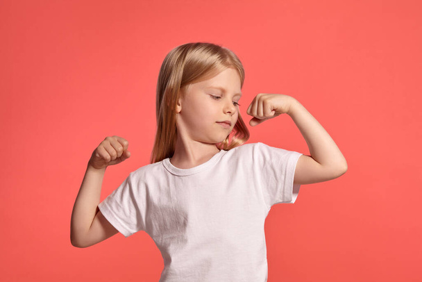 Close-up studio shot of a beautiful blonde little female with a long hair, in a white t-shirt, showing her muscles and looking at them while posing over a pink background with copy space. Concept of a - Foto, afbeelding