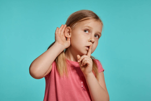 Close-up shot of a nice blonde little lady with two ponytails on her head, in a pink dress, acting like listening to someone while posing against a blue background with copy space. Concept of a joyful - Foto, afbeelding