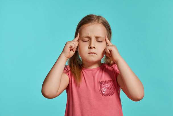 Close-up shot of pretty blonde child with two ponytails on her head, in a pink dress, thinking about something while posing against a blue background with copy space. Concept of a joyful childhood. - Foto, imagen