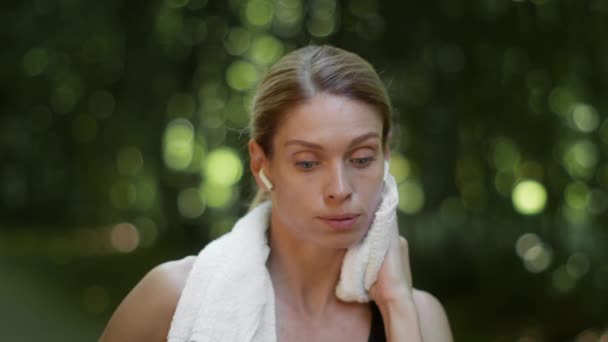 Exhausting sport training. Close up portrait of tired woman athlete wearing earbuds wiping her face with towel, feeling overworked after workout outdoor, slow motion - Video