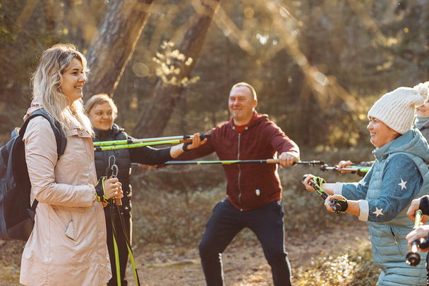 Smiling woman coach, sports trainer with group of people teach Scandinavian, Nordic walking squatting exercises with sticks in forest, nature. Exercises hike education senior people. Leisure activity - Фото, изображение