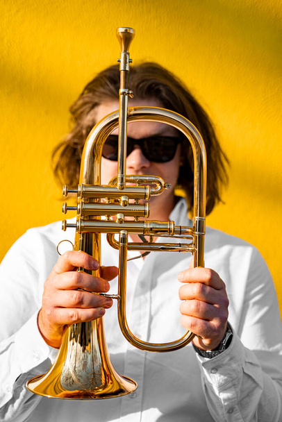 Portrait of young caucasian long-haired male musician jazzman in white shirt and sunglasses holding golden trumpet in front of himself, standing near yellow wall on street outside on sunny day - Photo, Image