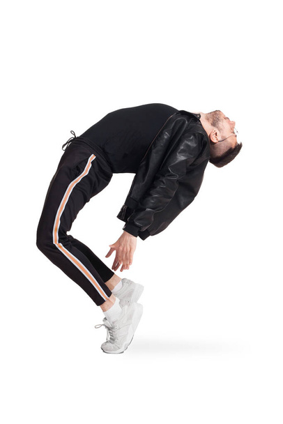 Full-length photo of a modern performer in glasses, black leather jacket, t-shirt, sports pants and light sneakers fooling around in studio. Indoor photo of a funny man dancing tilting back isolated - Photo, image
